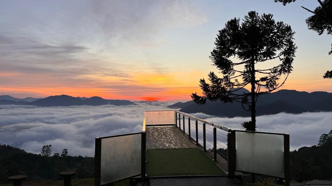 Pôr do sol sobre um mar de nuvens visto de uma plataforma, com montanhas e árvores em silhueta. Tons de laranja e dourado.