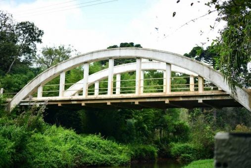 Ponte arqueada branca sobre um rio, rodeada por folhagem verde.
