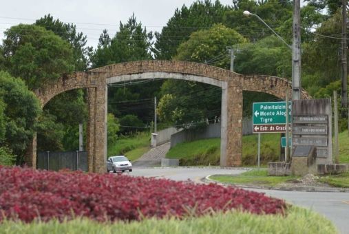 Entrada com arco de pedra, sinalização, um carro passando e flores vermelhas.
