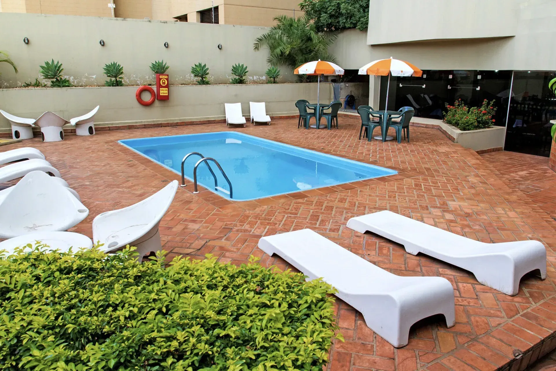 Área da piscina com espreguiçadeiras, mesas com guarda-sóis e um pátio de tijolos.