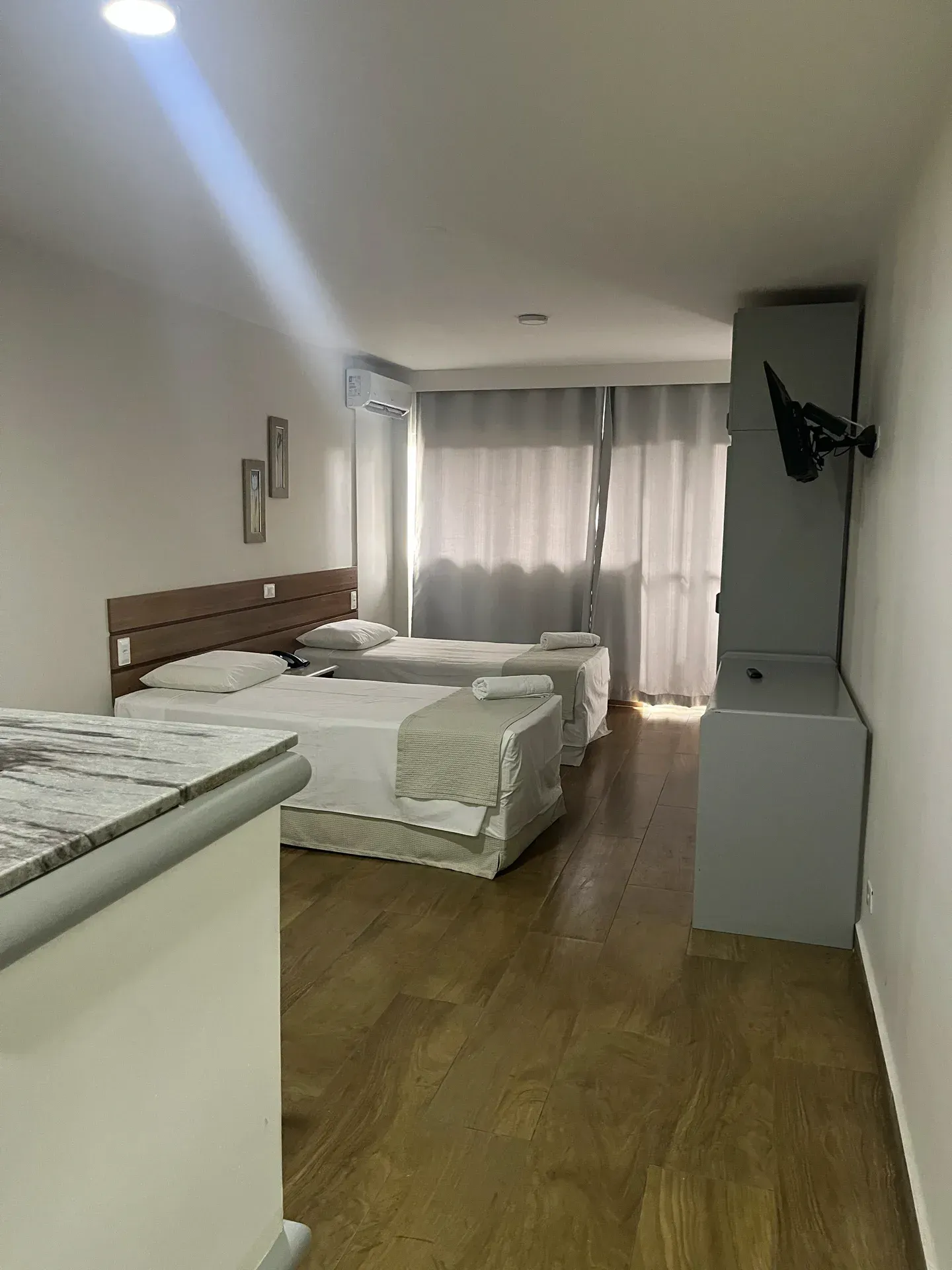 Quarto com duas camas de solteiro, piso de madeira e um armário cinza com uma TV fixada na parede.