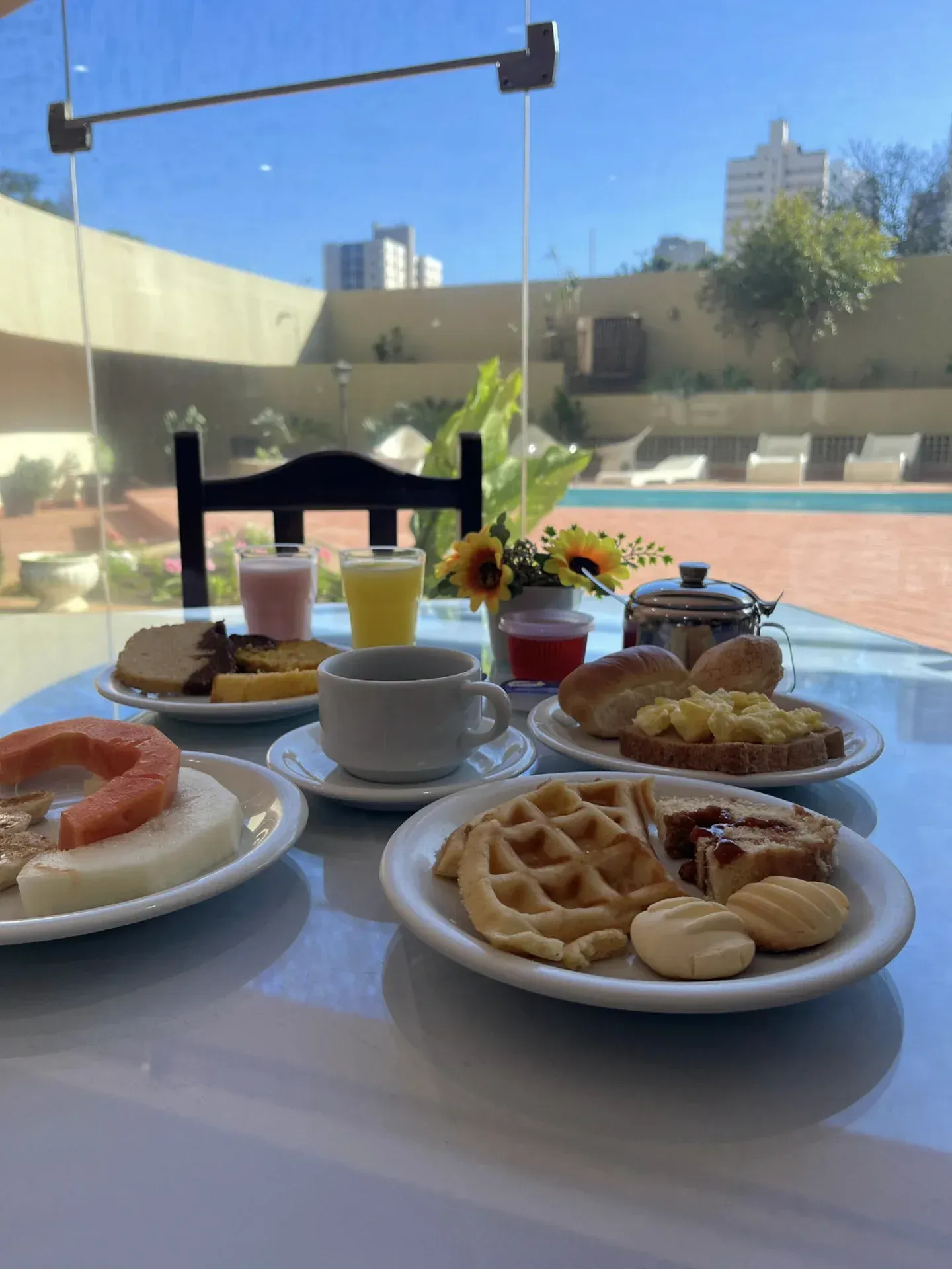 Café da manhã servido em uma mesa, incluindo waffles, frutas e bebidas, com vista para a piscina através de uma janela.