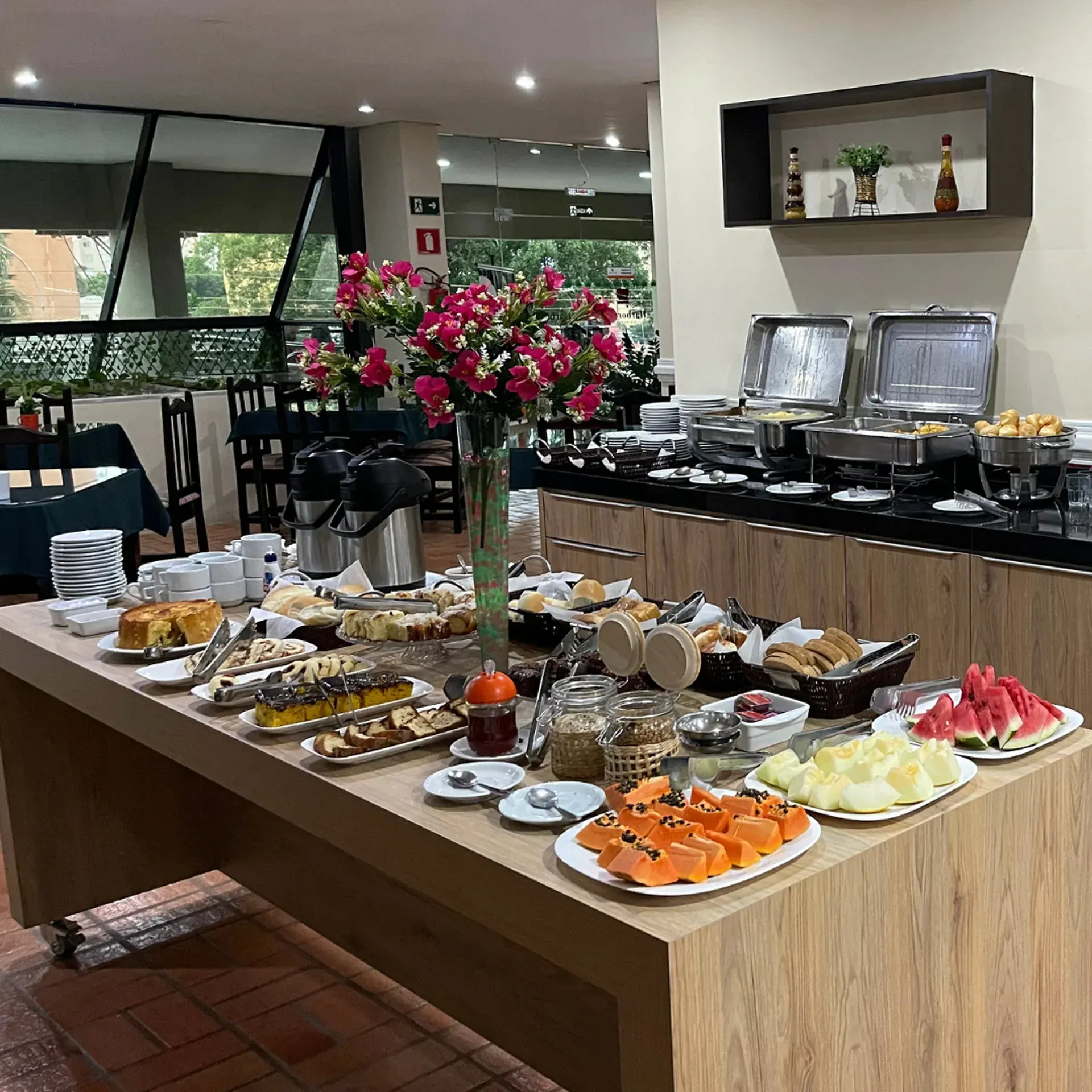 Buffet de café da manhã com diversas opções de comida em um balcão de madeira, em um ambiente de restaurante.