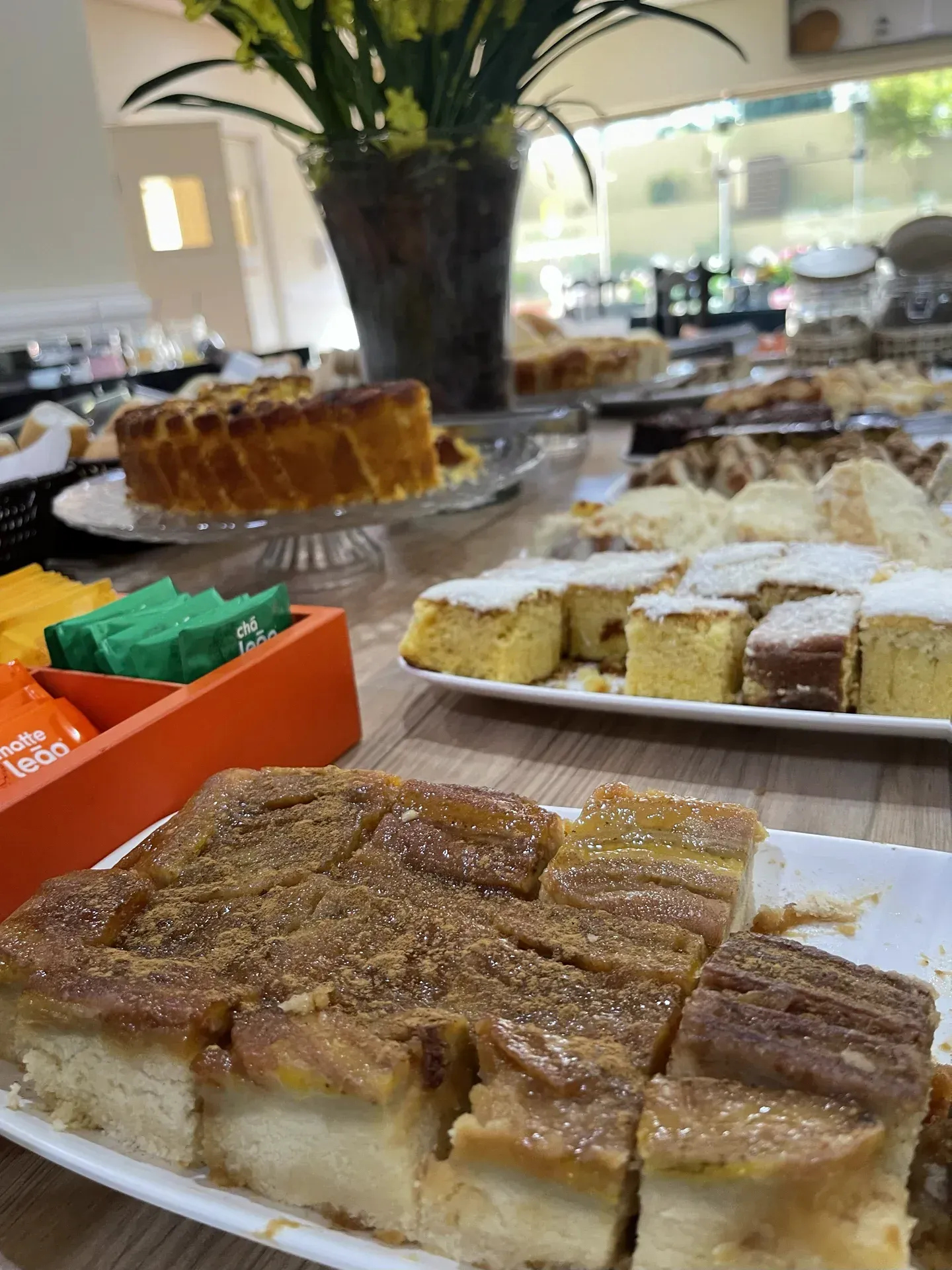 Buffet de sobremesas com diversos bolos e doces, sobre uma mesa de madeira, com saquinhos de chá em uma lata laranja.