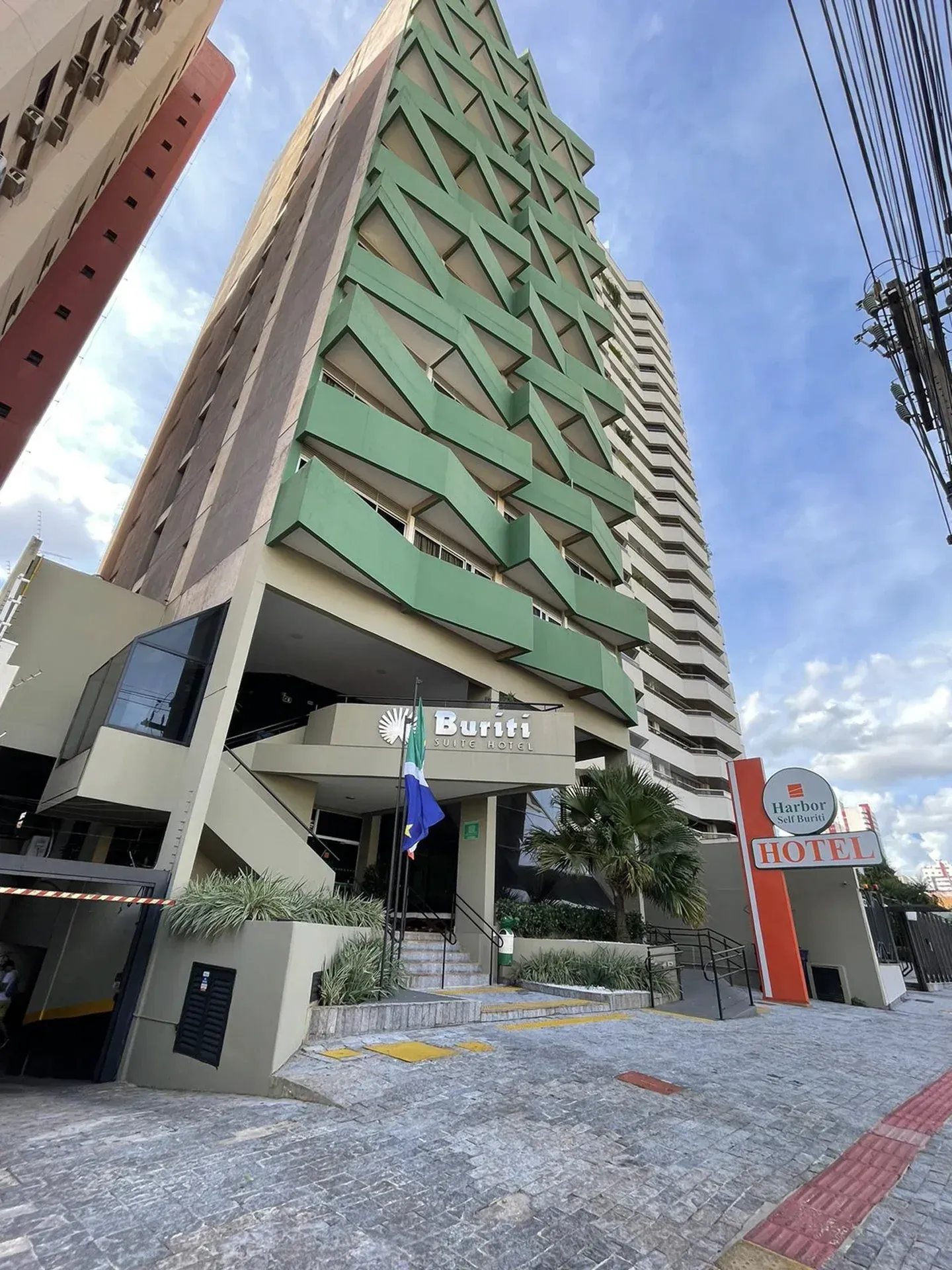 Edifício moderno de hotel de vários andares com fachada verde e bege; escadaria de entrada, placa, bandeira azul, céu limpo.