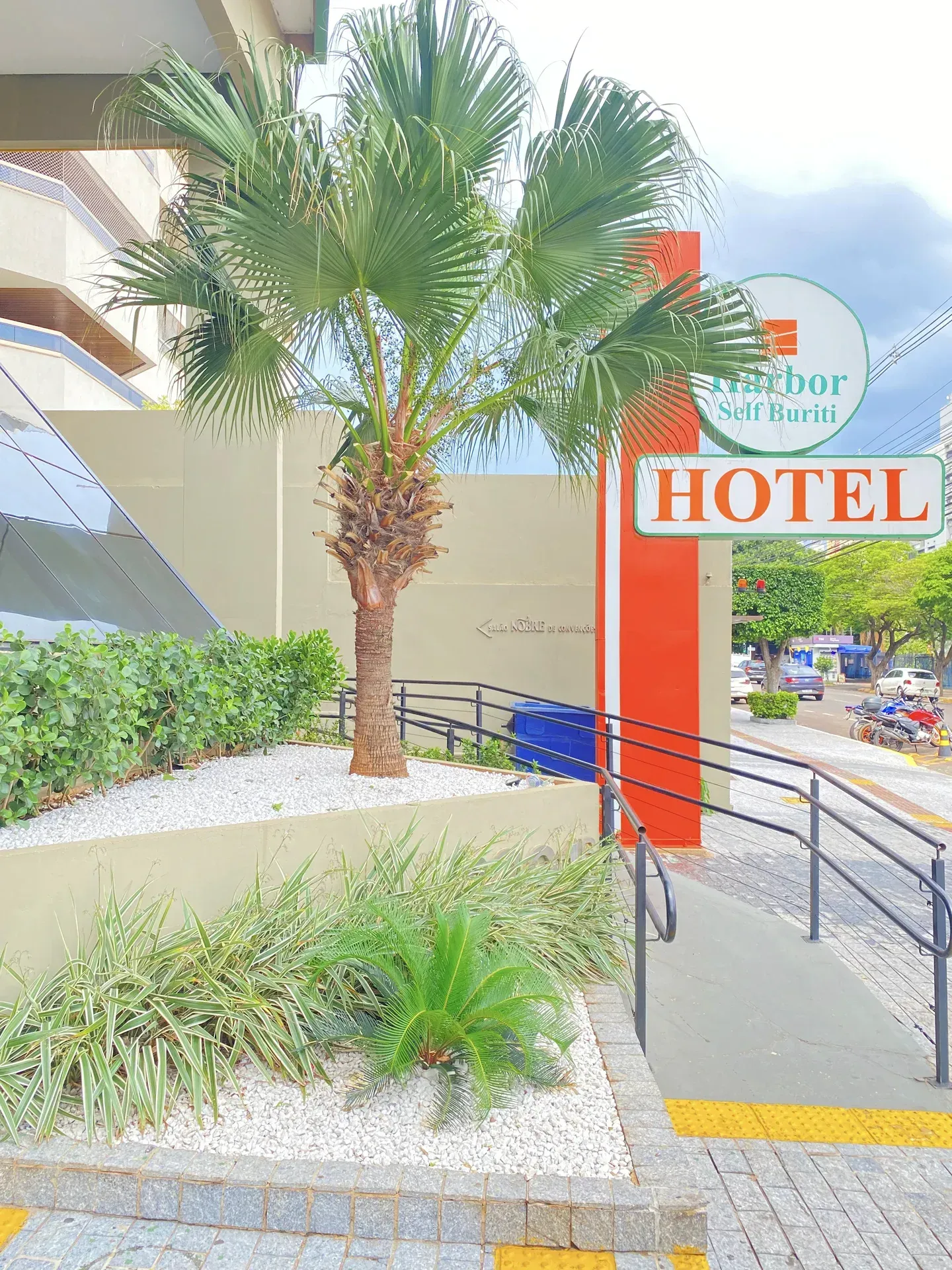 Entrada do hotel com palmeira, placa, rampa de acesso e paisagismo.