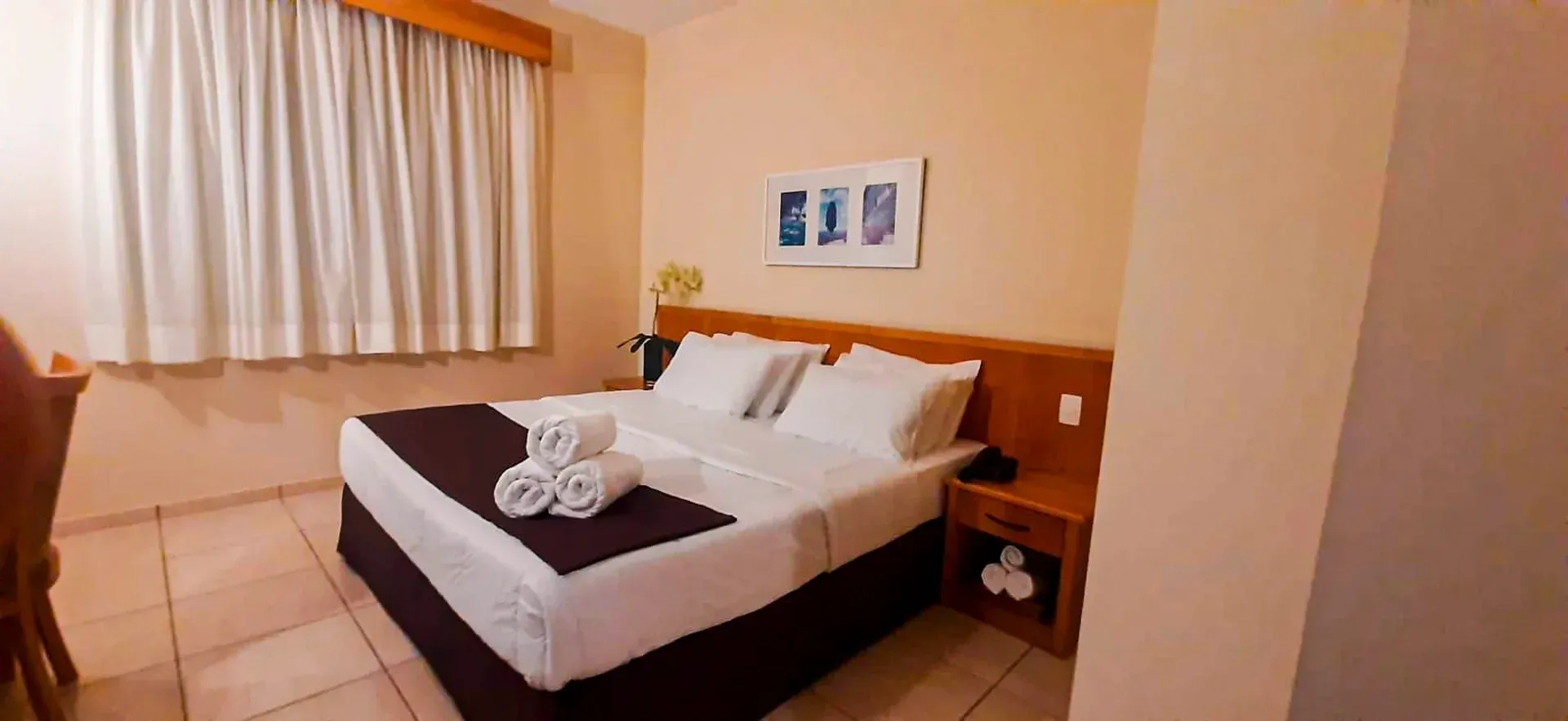 Quarto de hotel com cama, mesa de cabeceira, janela e obras de arte na parede.