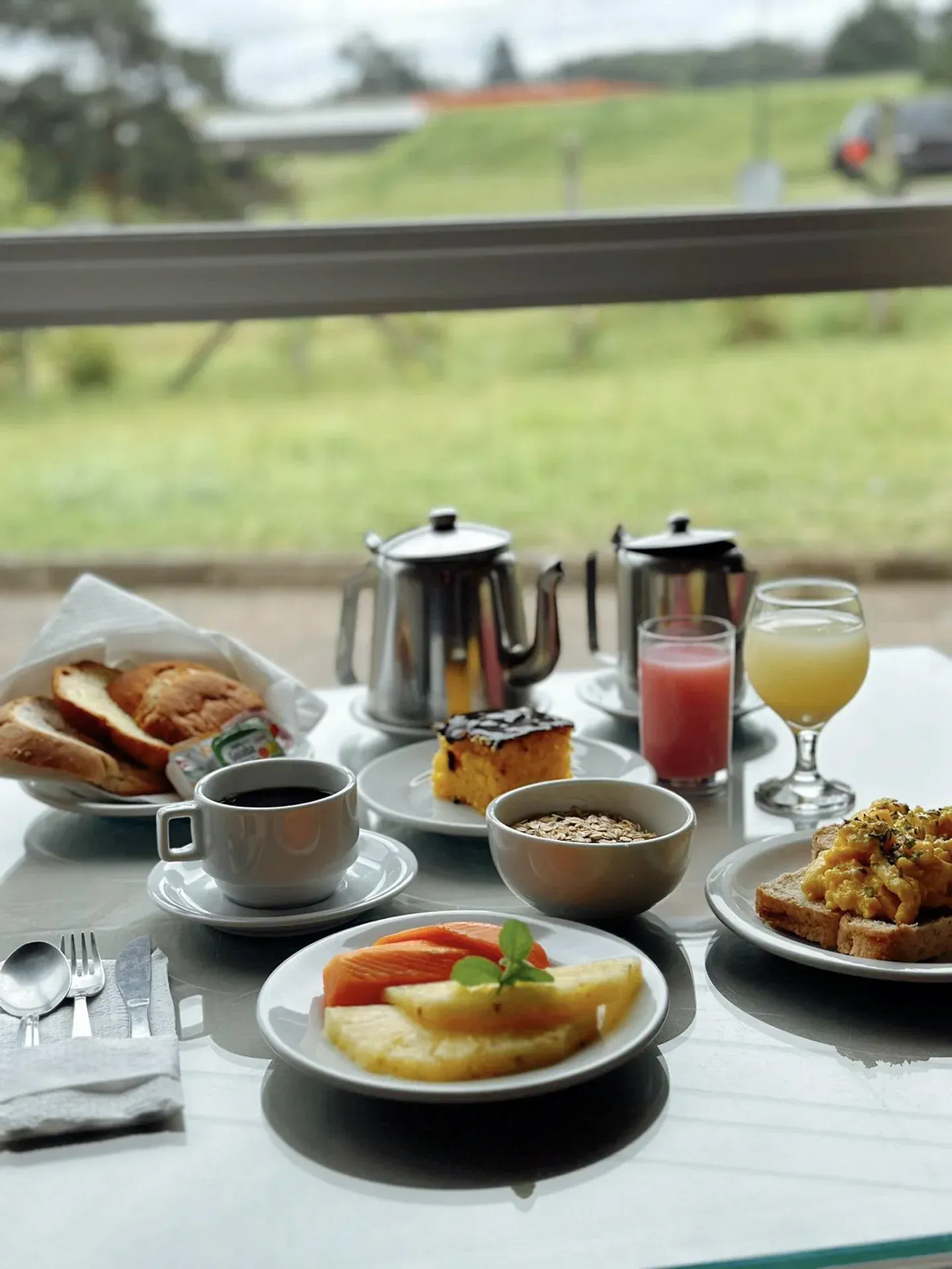 Café da manhã servido em uma mesa com frutas, pão, cereais, café e suco, com vista para um jardim verdejante.