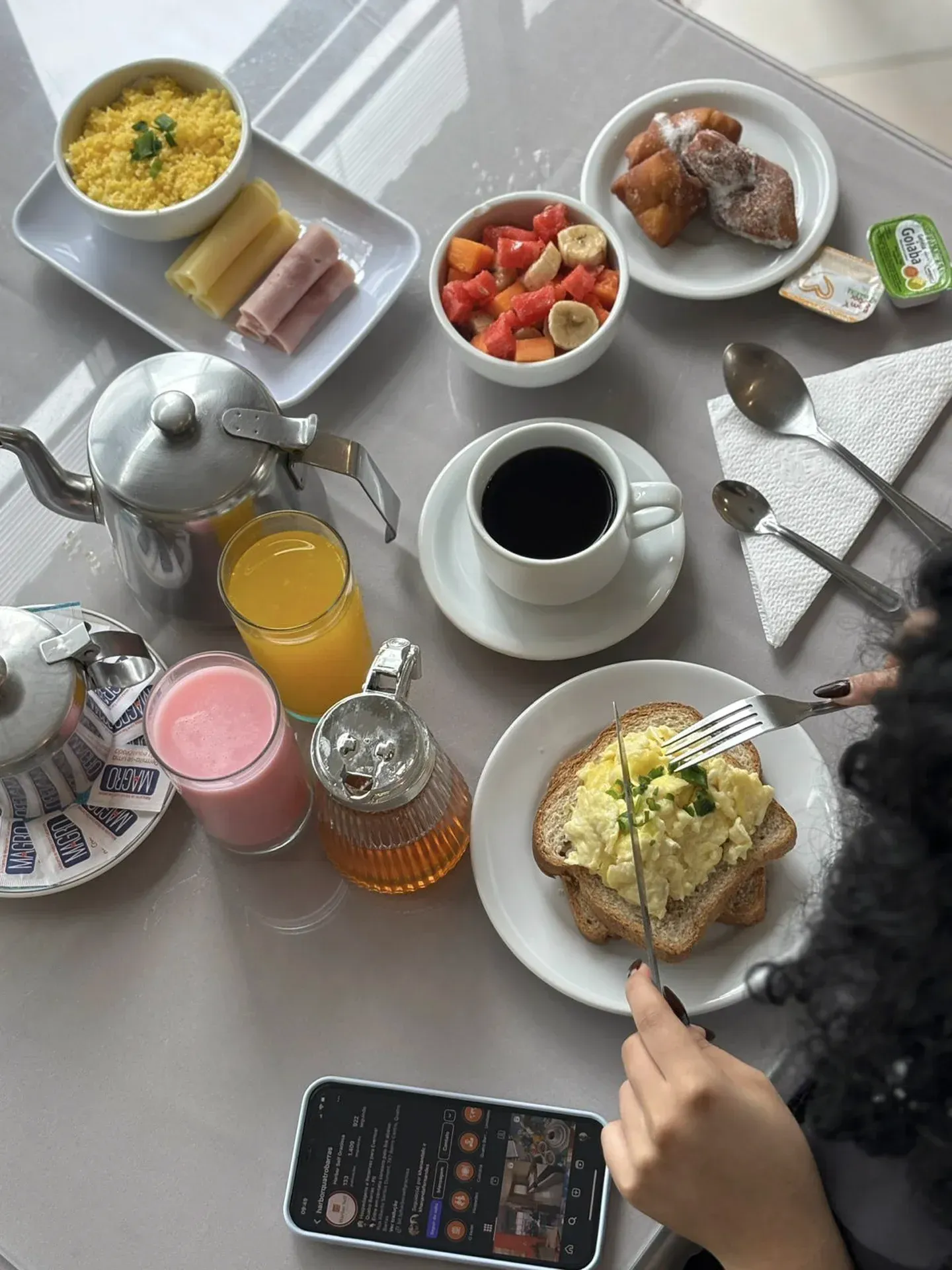 Café da manhã: ovos mexidos, café, suco, frutas, doces, presunto e uma pessoa comendo.