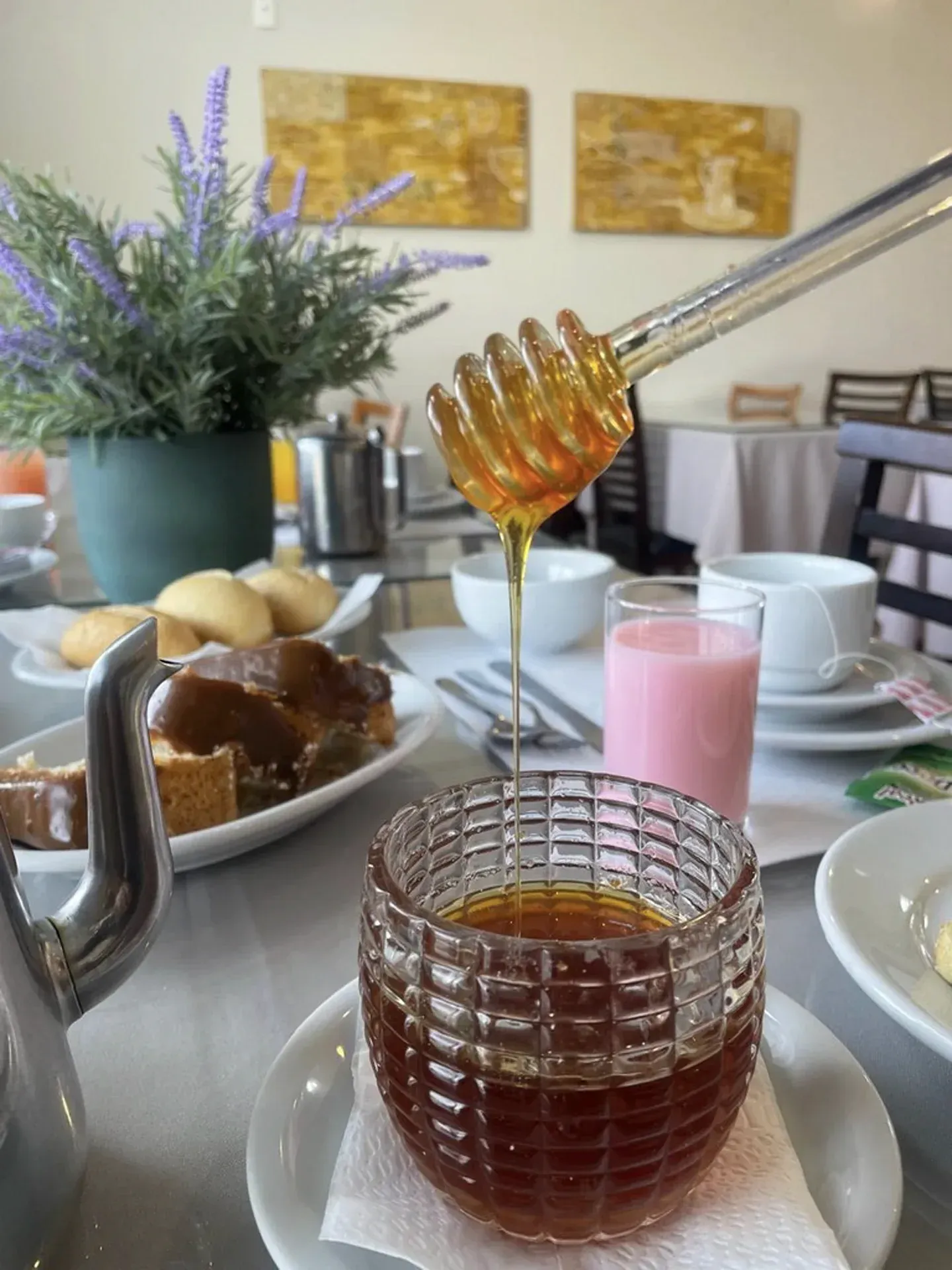 Mel escorrendo de uma concha para um pote de cristal, parte de um café da manhã servido com torradas, pãezinhos e uma bebida rosa.