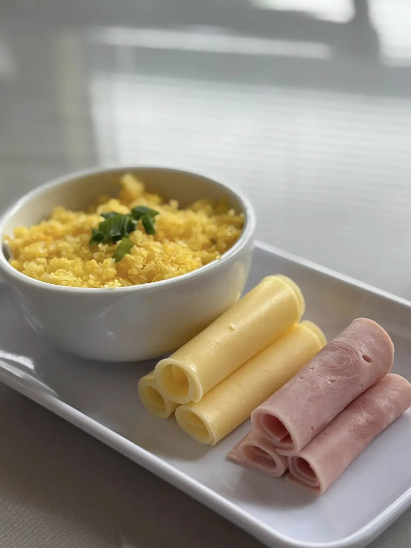 Tigela de cuscuz amarelo com rolinhos de presunto e queijo em um prato retangular branco.