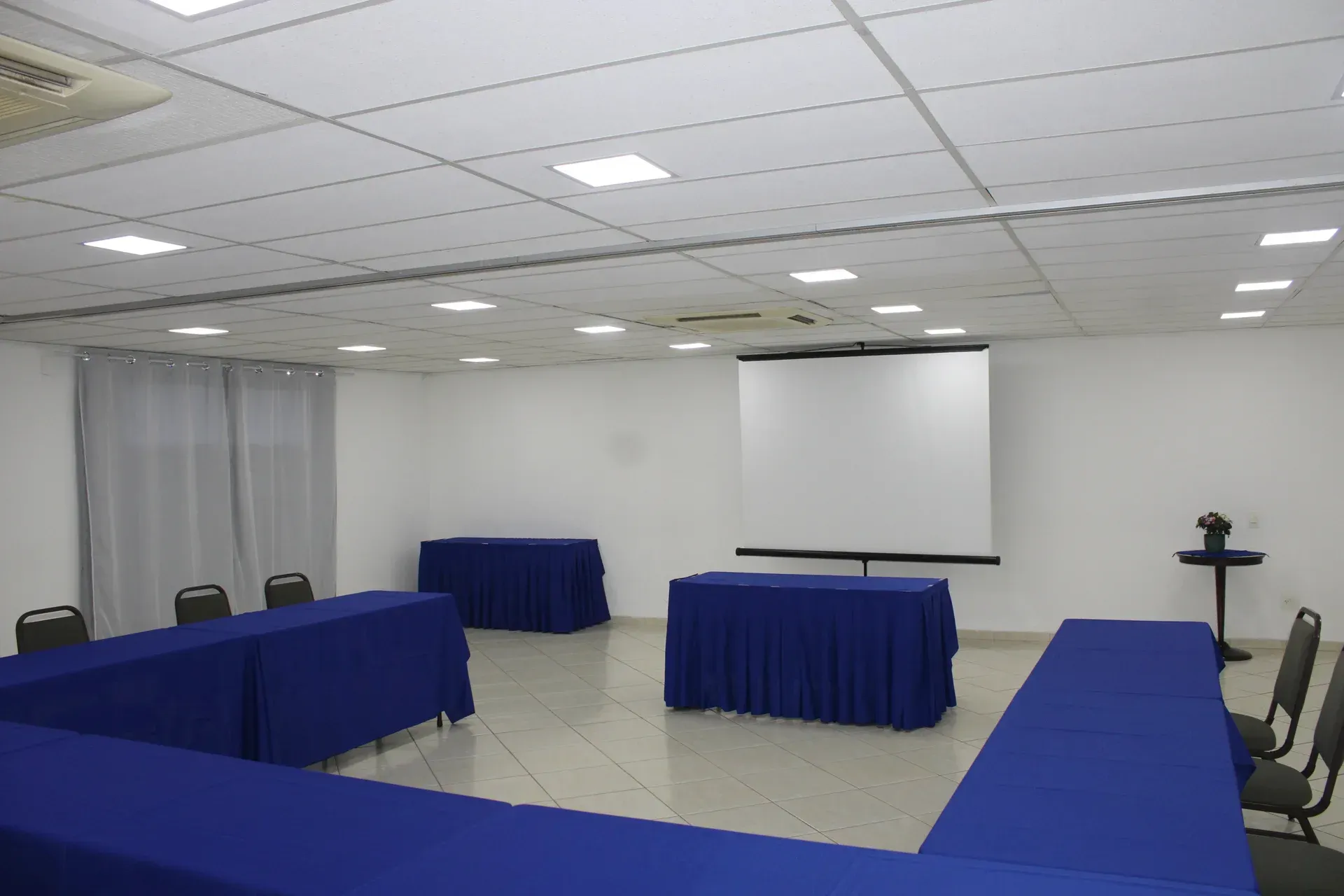 Sala de conferências com toalhas de mesa e cadeiras azuis, tela de projeção e paredes brancas.