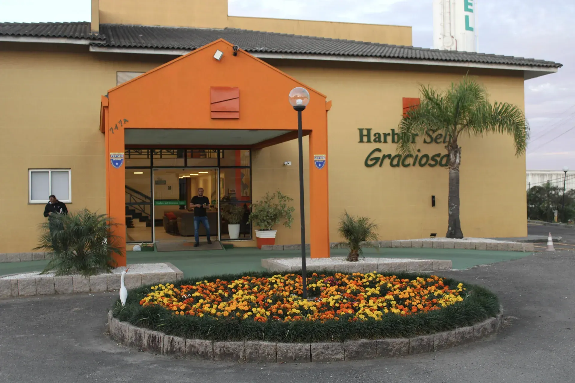 Exterior do hotel Harbor Self Gracioso, entrada com toldo laranja e canteiro de flores em frente.