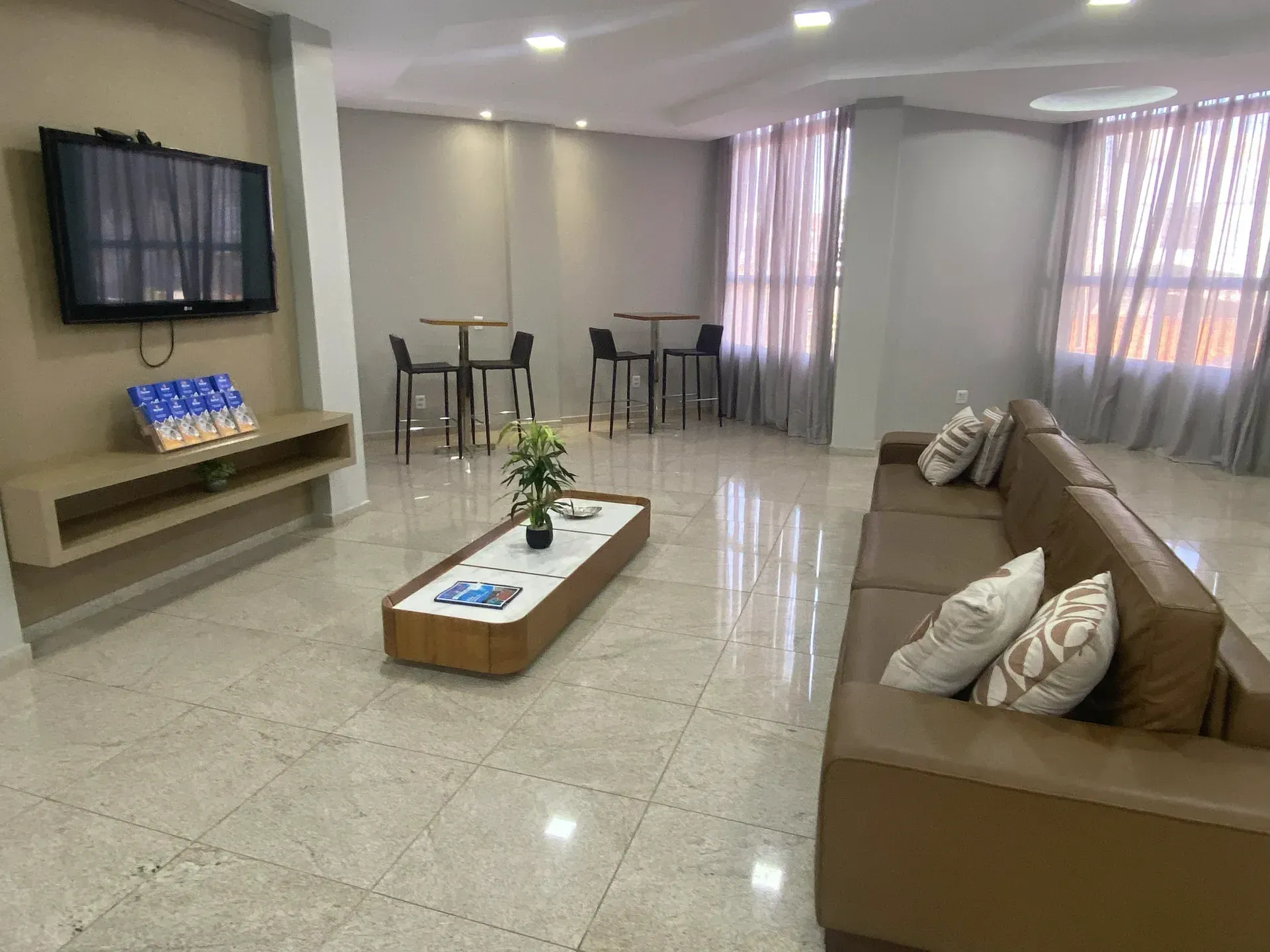 Área de estar com sofá marrom, mesa de centro, TV, mesas e cadeiras altas de bar e janela com cortinas.