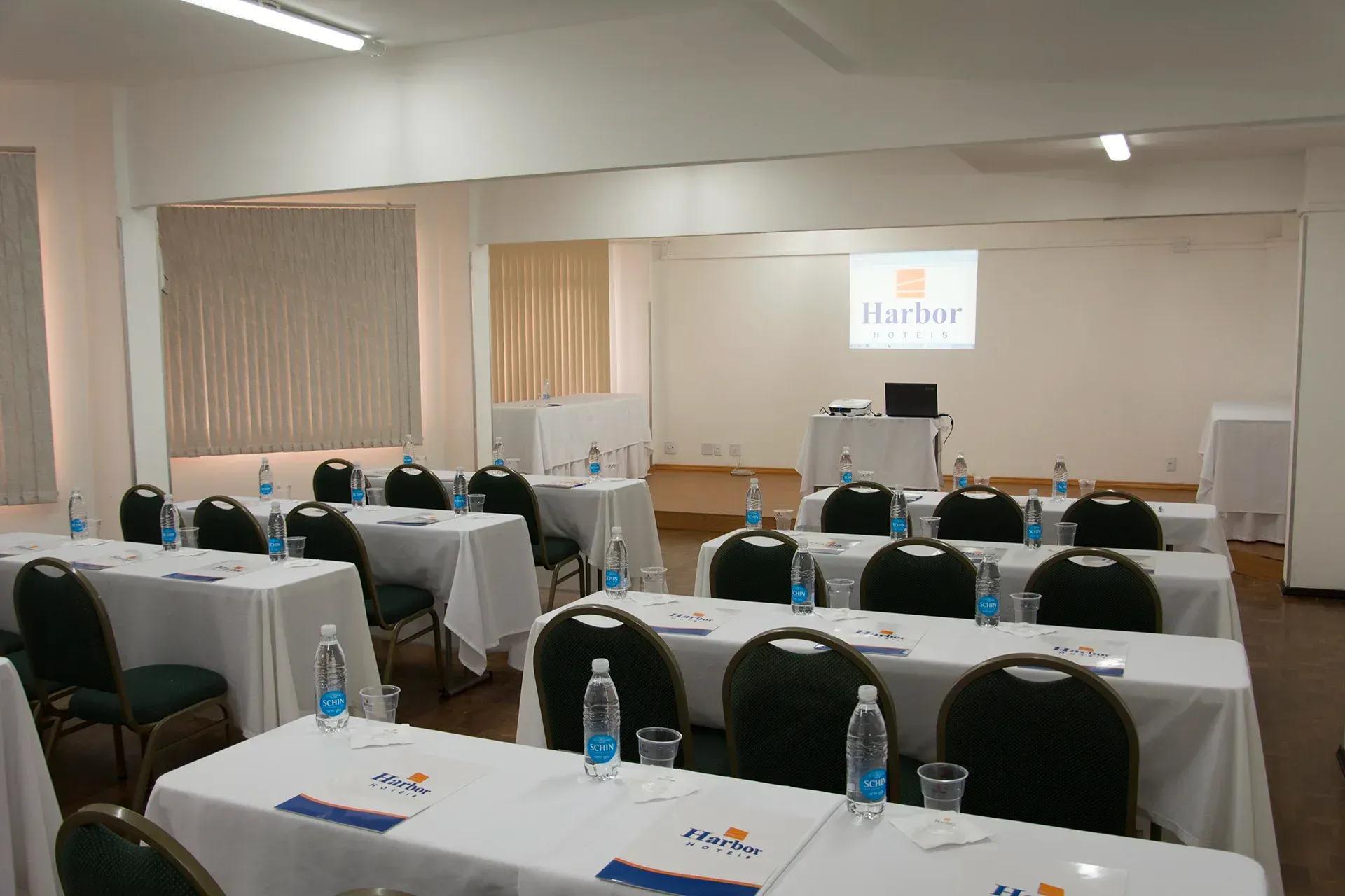 Sala de conferências equipada com mesas, cadeiras e tela de projeção exibindo um logotipo.