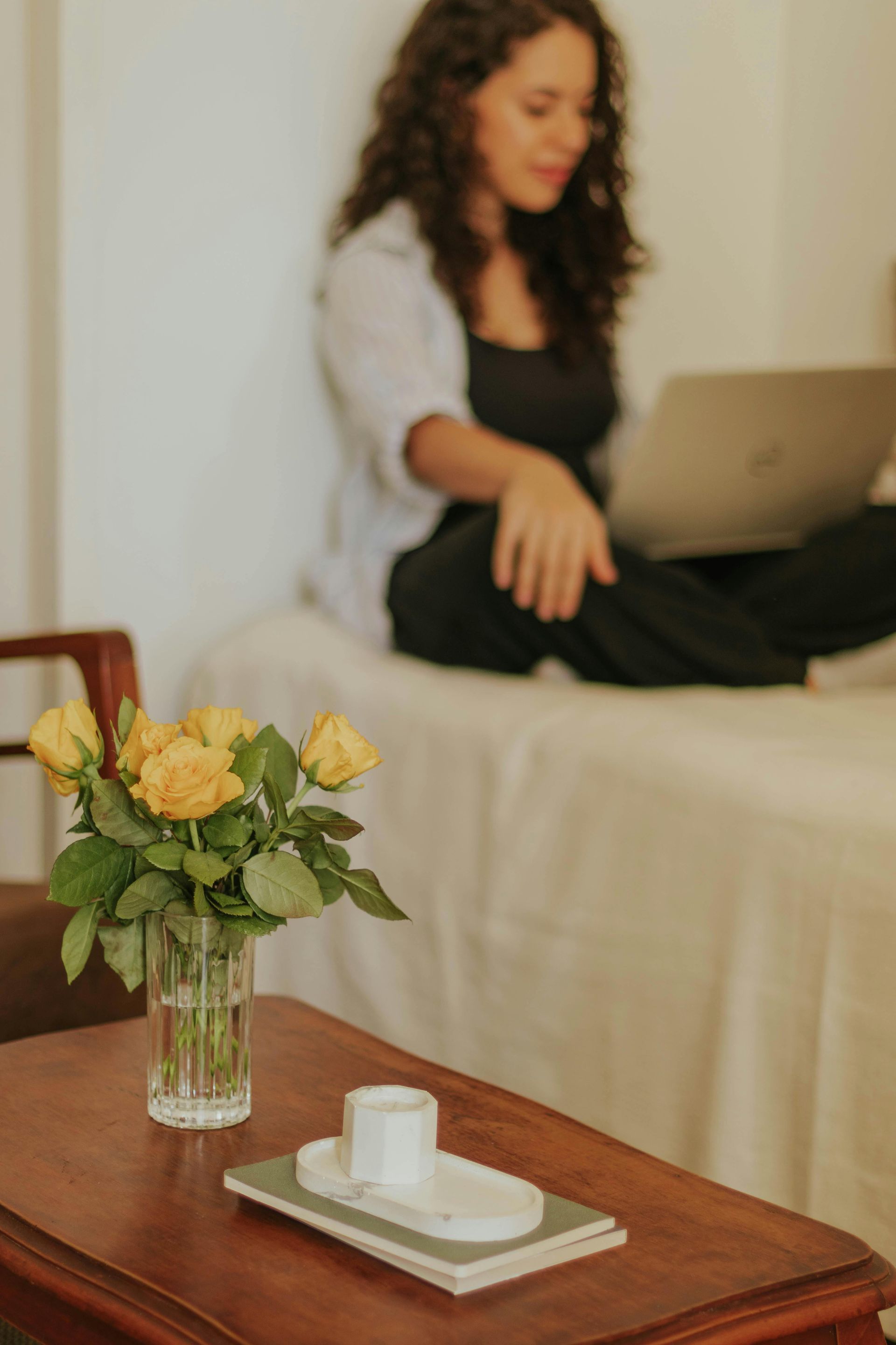 Rosas amarelas num vaso sobre uma mesa; ao fundo, uma pessoa deitada na cama trabalha num computador portátil.
