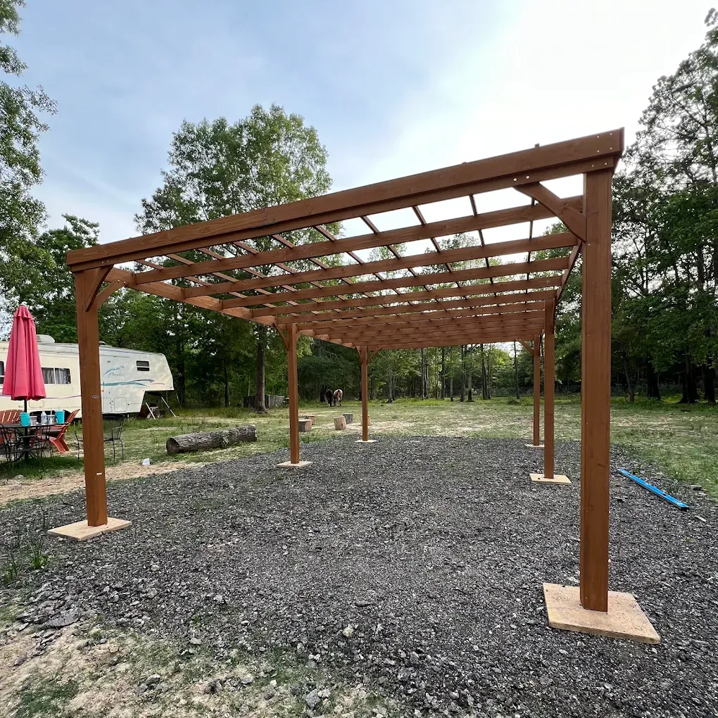 Brown wooden pergola frame on gravel in a park setting with trees and a picnic table nearby