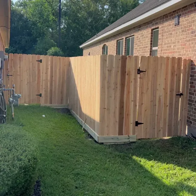 Wooden fenced enclosure beside a brick house in a grassy backyard, with a gate and utility meter visible.