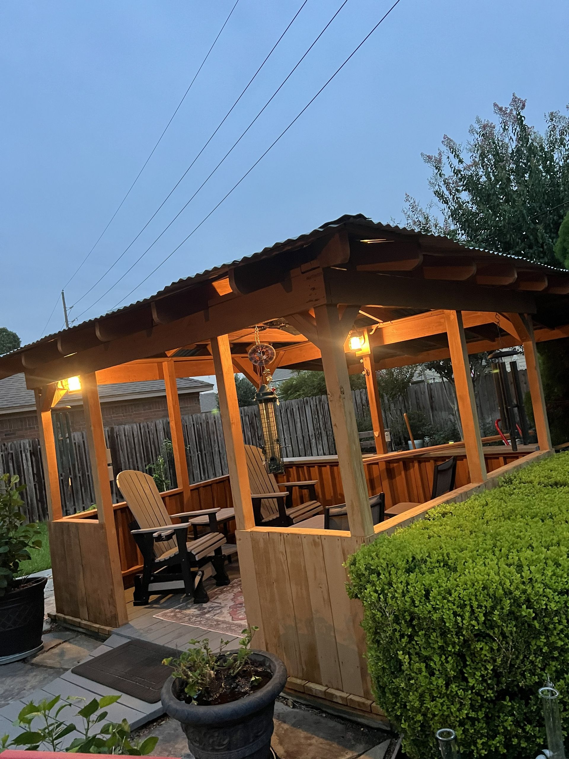 Wooden outdoor pavilion with warm lights, benches, and a hedge at dusk