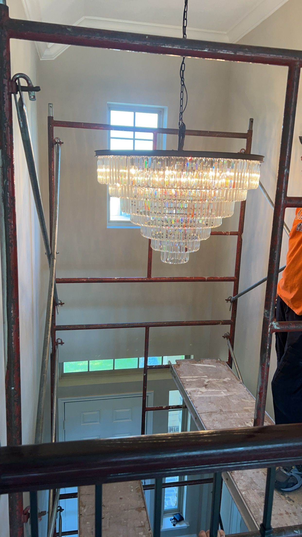 Crystal chandelier hanging in a tall stairwell with dark railings and a bright window above.