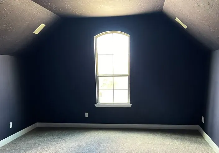 Empty dark blue attic room with a centered arched window and gray carpet.
