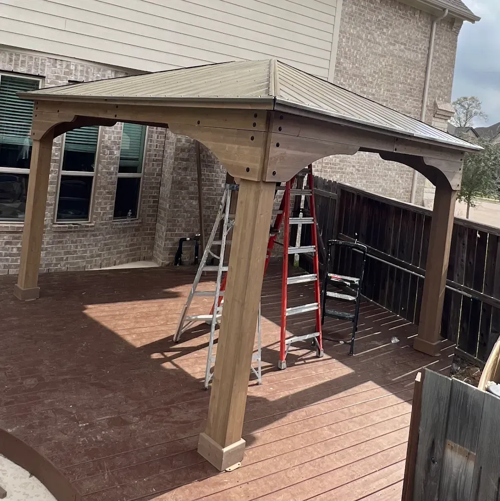 Backyard wooden pergola under construction with ladders on a brick patio and deck.