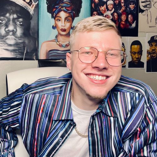 Smiling person in glasses and a striped shirt, seated before colorful portrait posters.