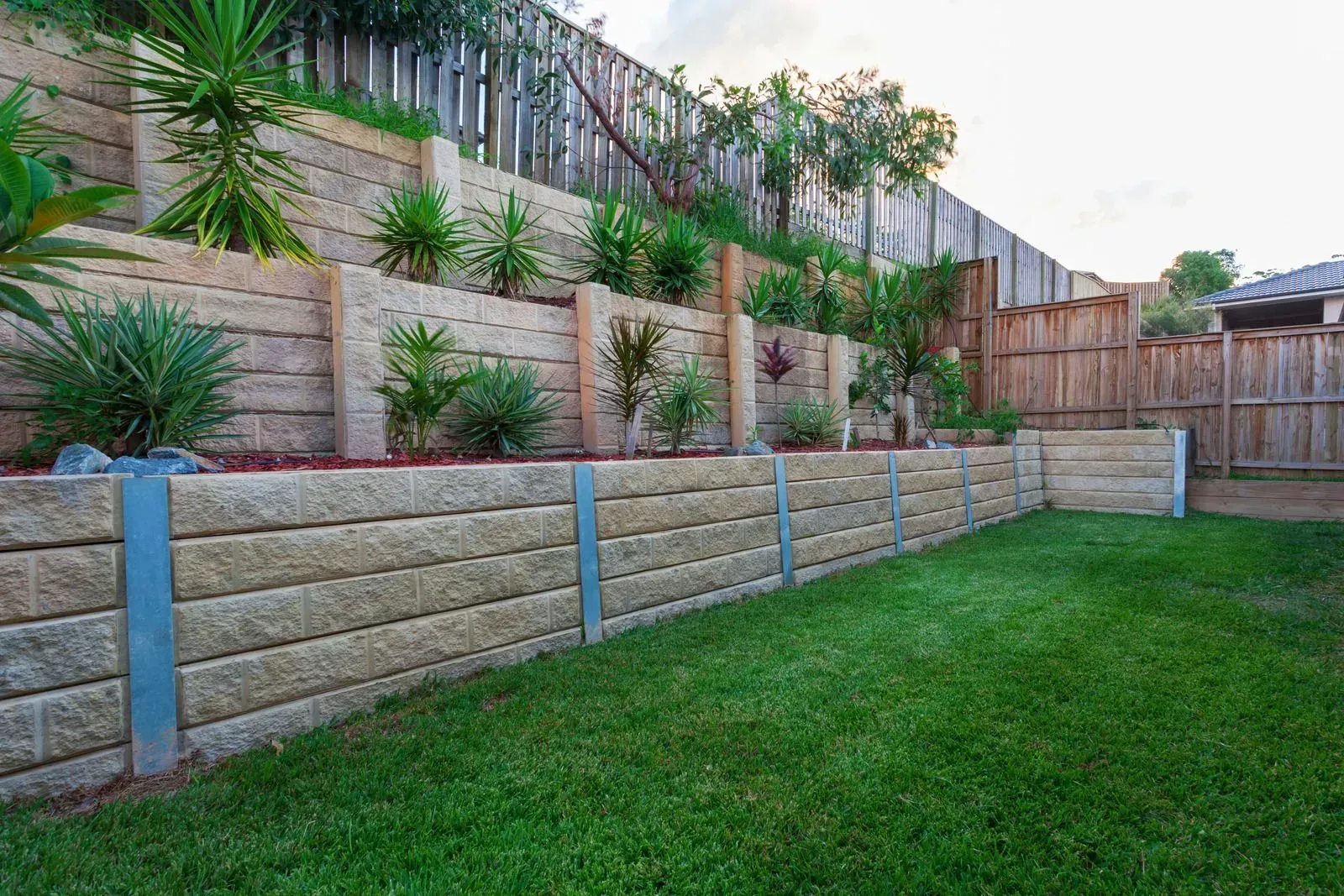 Retaining walls made of stacked stone with plants and grass, in a backyard setting with a wooden fence.
