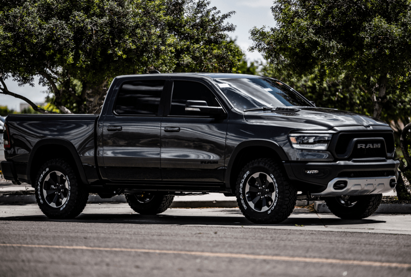 A dark gray RAM pickup truck parked on an asphalt street with trees in the background.