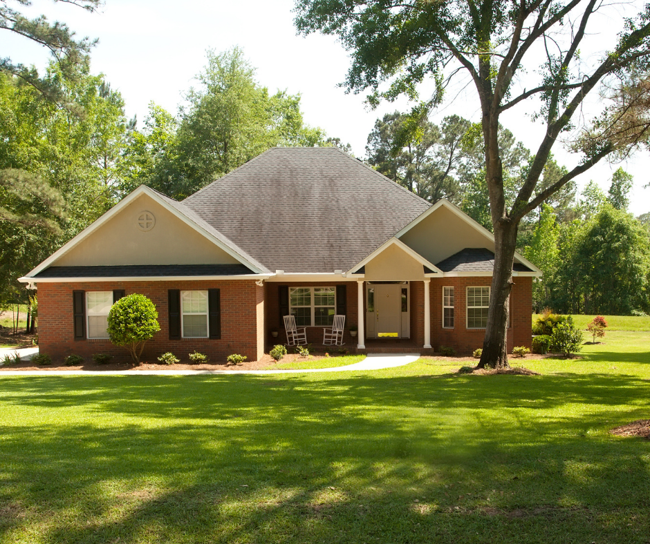 A single-story brick house with a dark shingled roof, set on a grassy lawn surrounded by trees on a sunny day.
