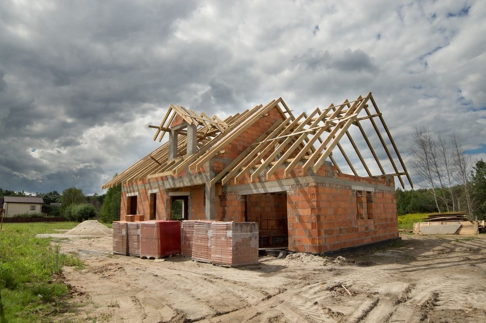 House under construction with brick walls, wooden roof frame, and cloudy sky.