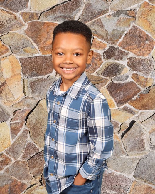 A young boy in a plaid shirt is smiling in front of a stone wall.