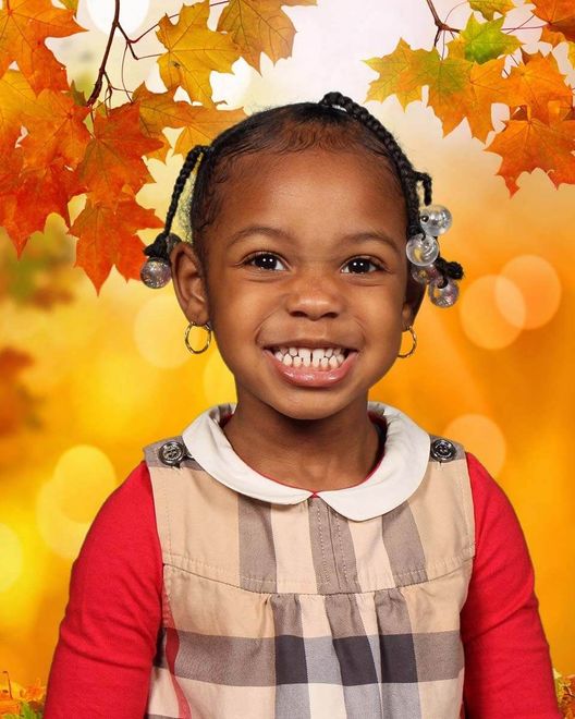 A little girl in a plaid dress is smiling in front of autumn leaves