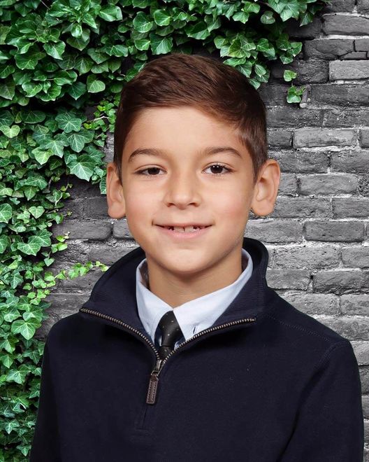 A young boy wearing a sweater and tie is smiling in front of a brick wall.
