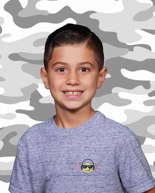 A young boy wearing a gray t-shirt with an emoji on it is smiling in front of a camouflage background.