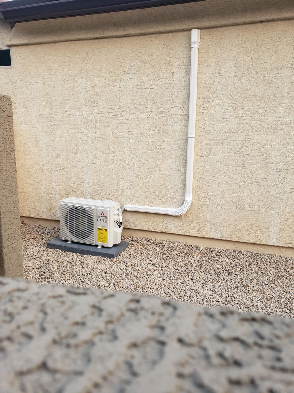 An outdoor air conditioning unit next to a stucco wall, with piping. Gravel ground.