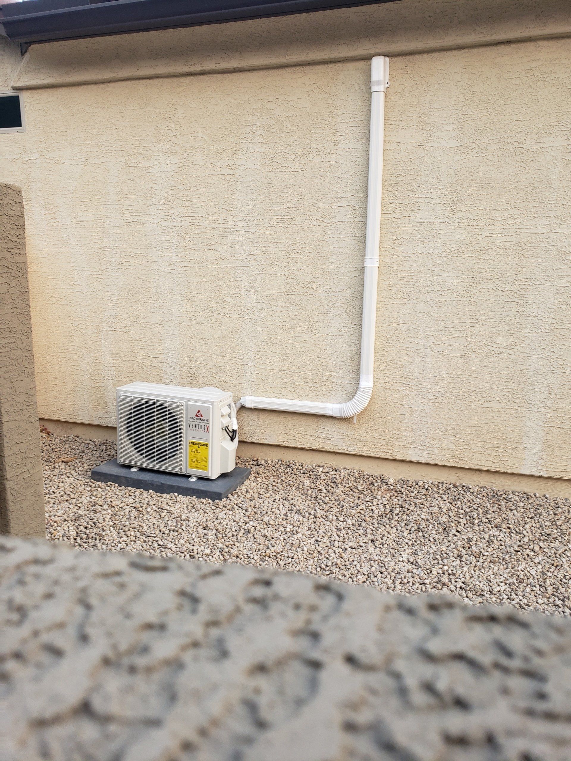 Air conditioning unit on a gravel bed against a stucco wall, with white conduit running upwards.