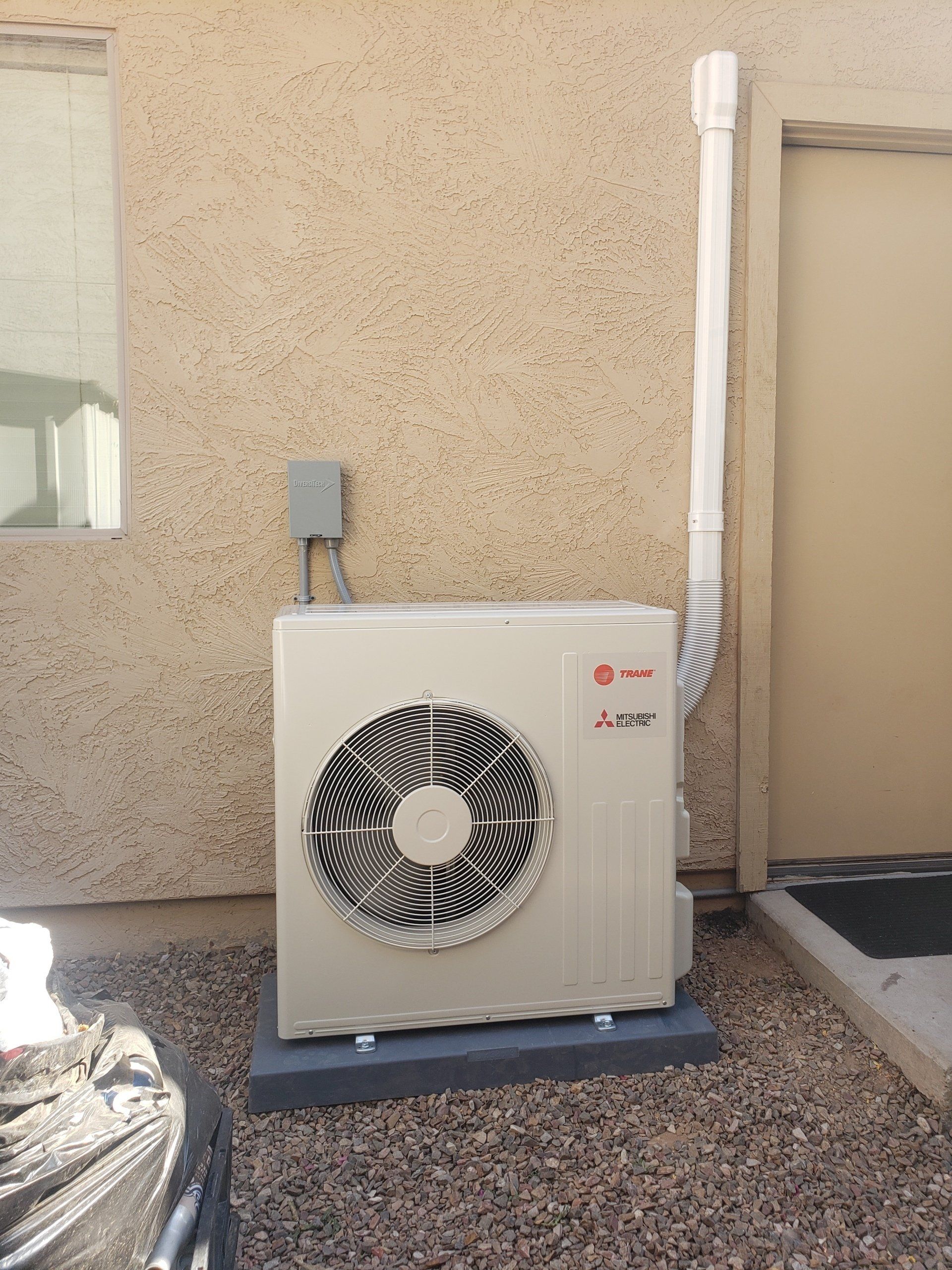 Outdoor air conditioning unit with white exterior, mounted on a concrete base, next to a building with a beige stucco wall.