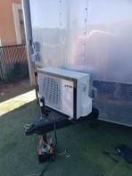 Air conditioner mounted on trailer hitch next to a silver trailer.