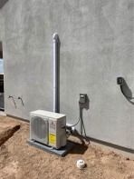 HVAC unit on a concrete pad against a light-gray stucco wall. Gray vent pipe and electrical box present. Dirt ground.
