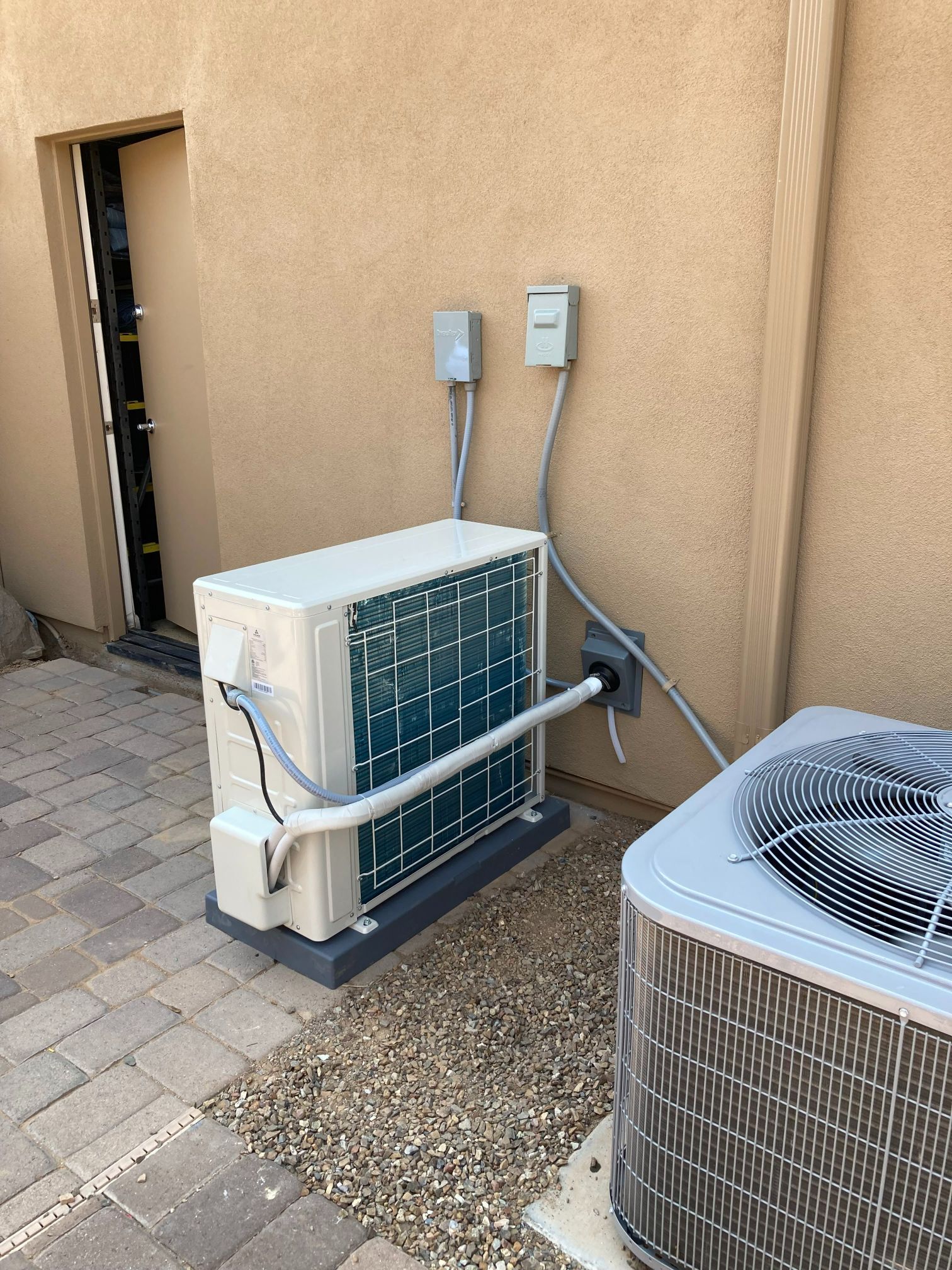 Outdoor HVAC units mounted on a patio; one is boxy, the other is a standard AC unit.