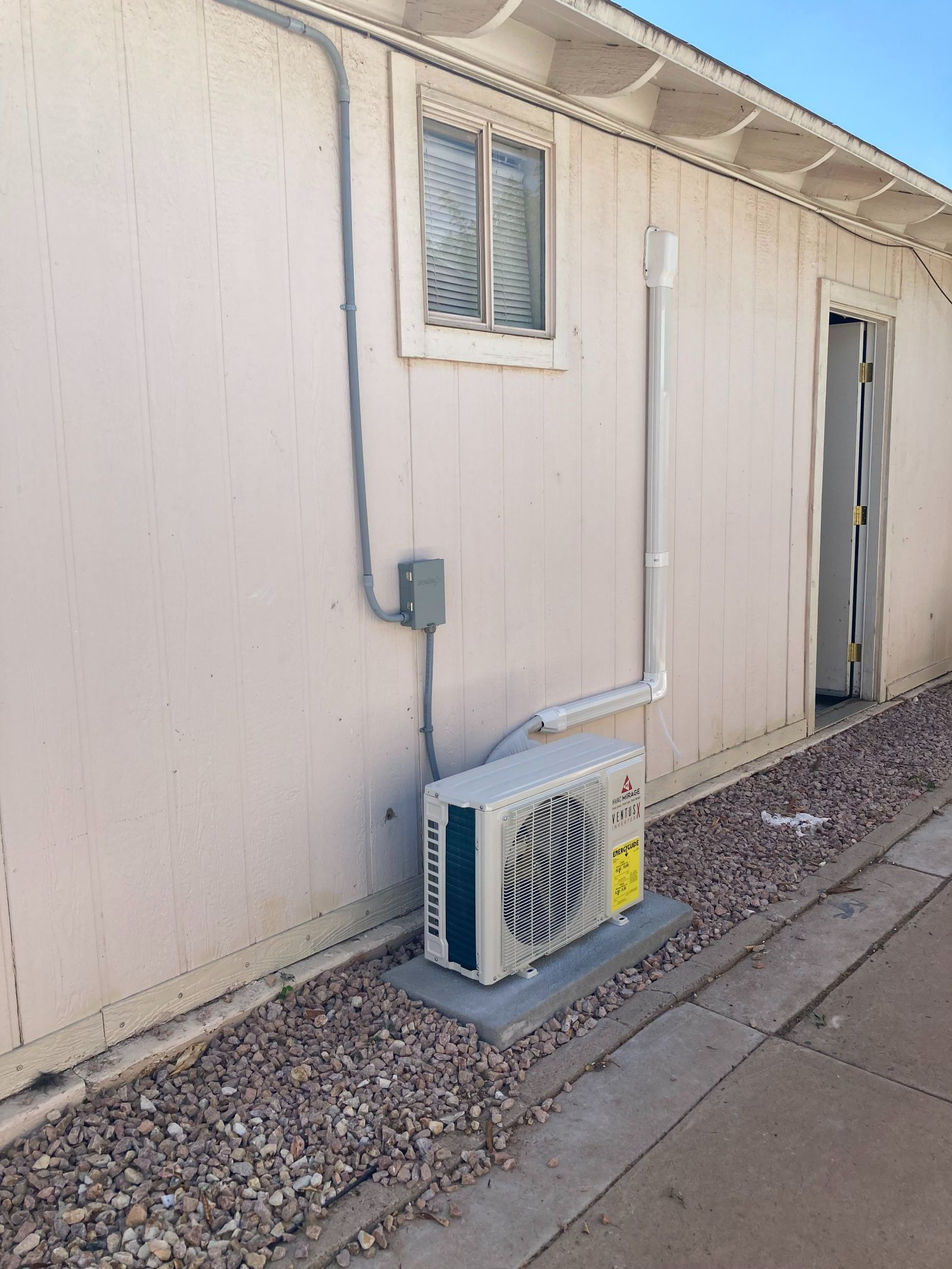 Outdoor air conditioning unit mounted on a concrete pad next to a building wall with a window and a door.
