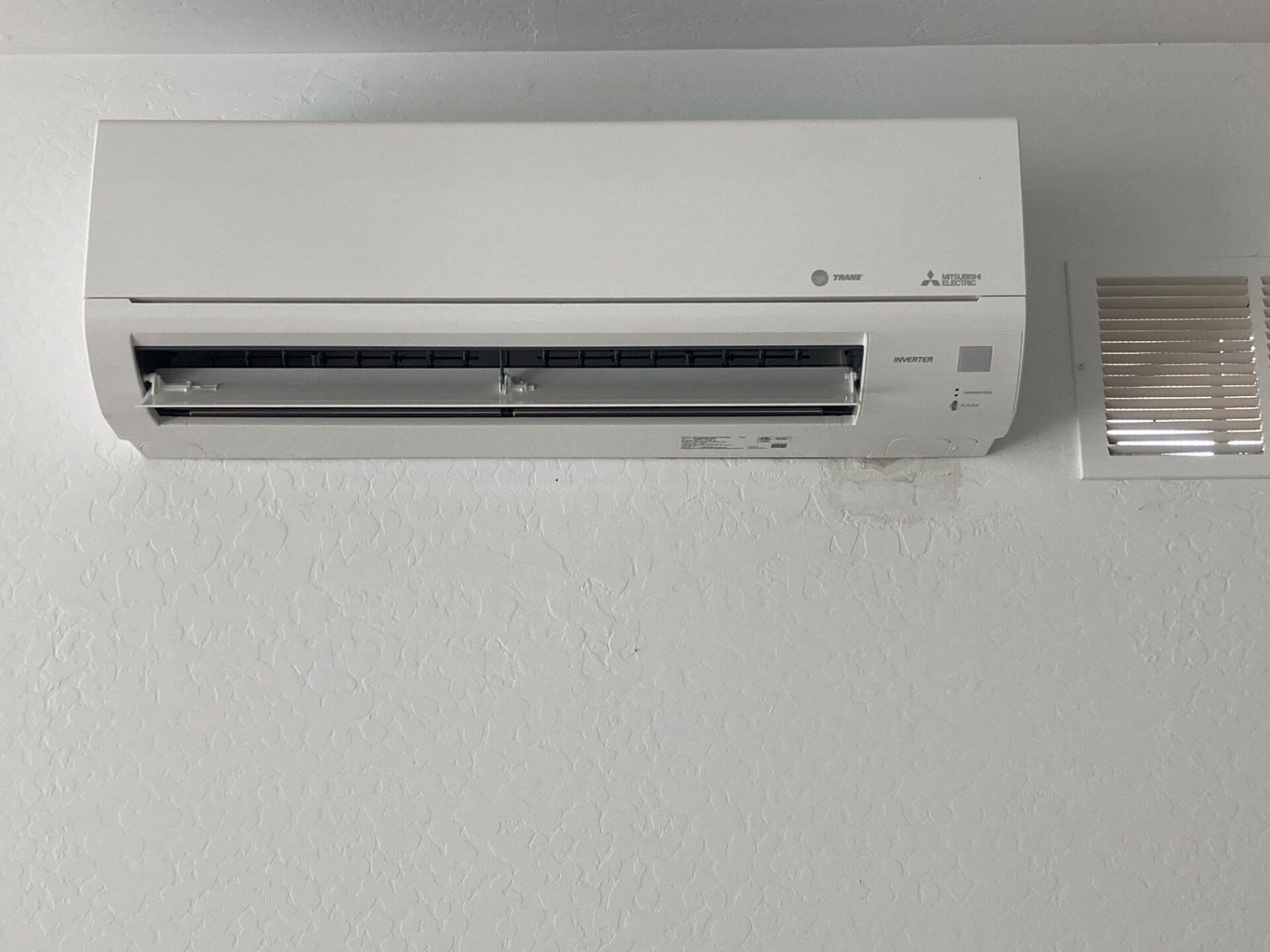 White wall-mounted air conditioner with a louvered vent, next to a small white square vent.