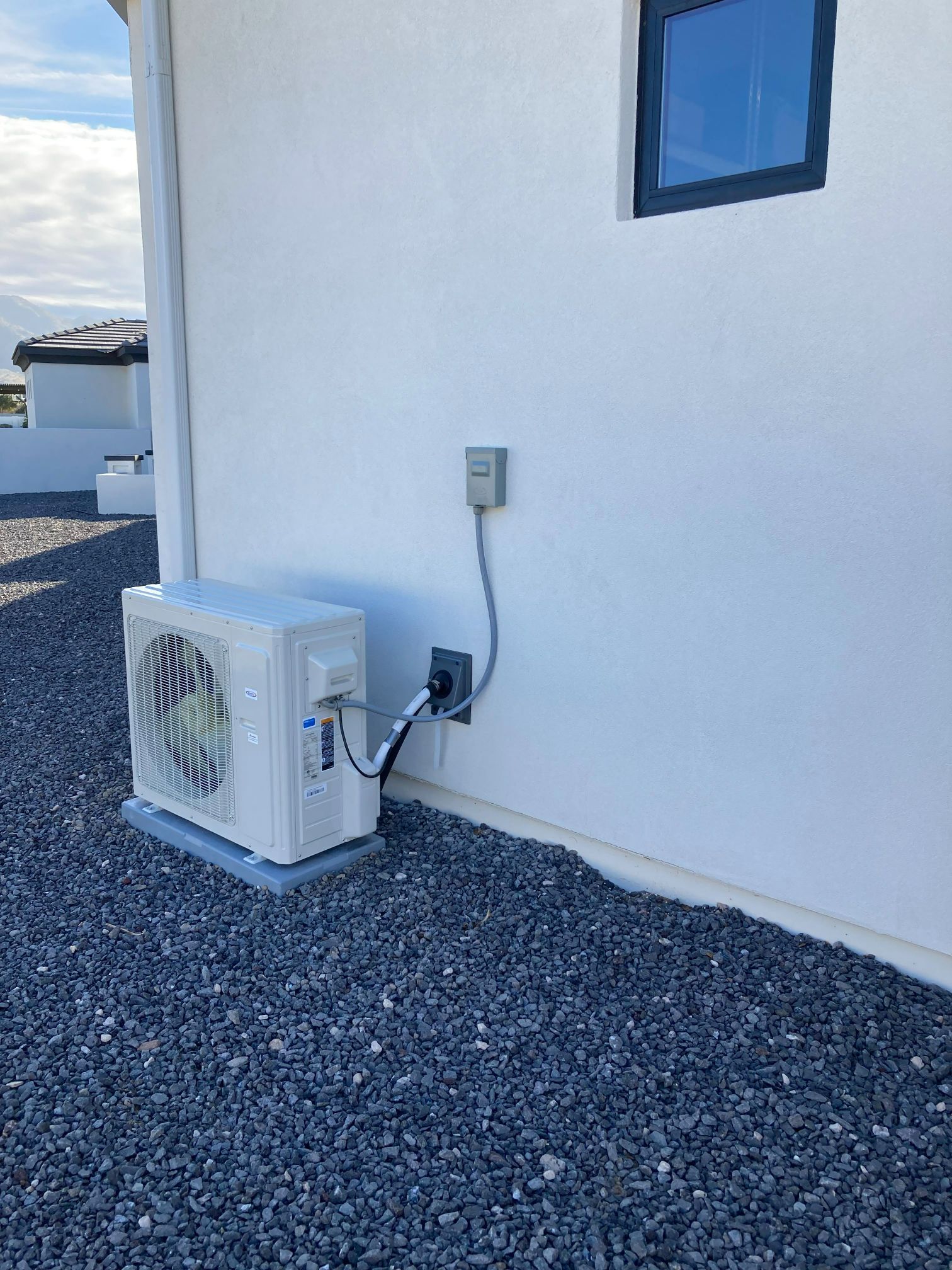 White air conditioner unit mounted on a gravel ground next to a white wall with a power outlet.