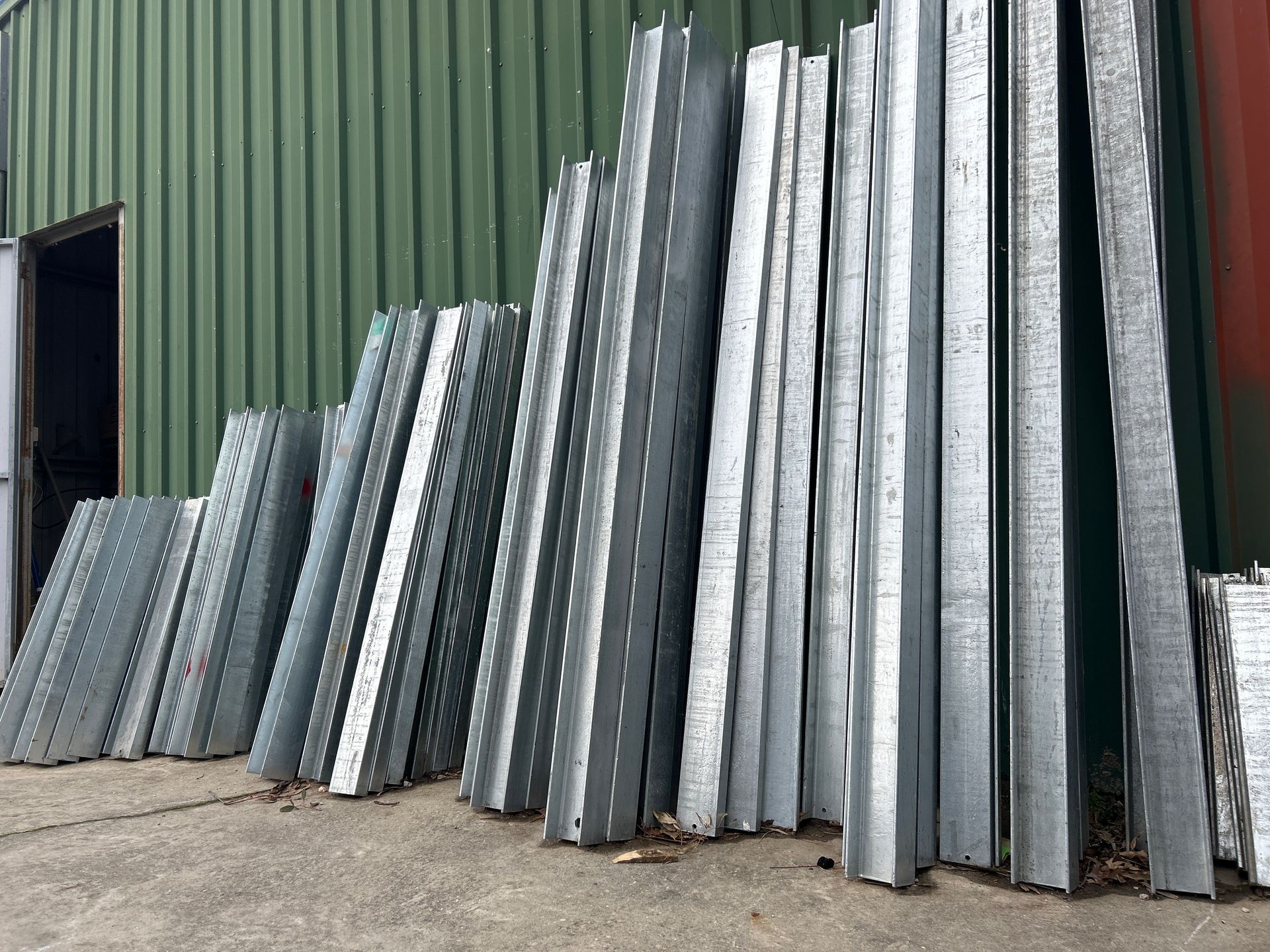 A Pile Of Metal Building Studs Leaning Against A Green Corrugated Metal Wall, In An Outdoor Setting — Wodonga Sand & Soil in Wodonga, VIC