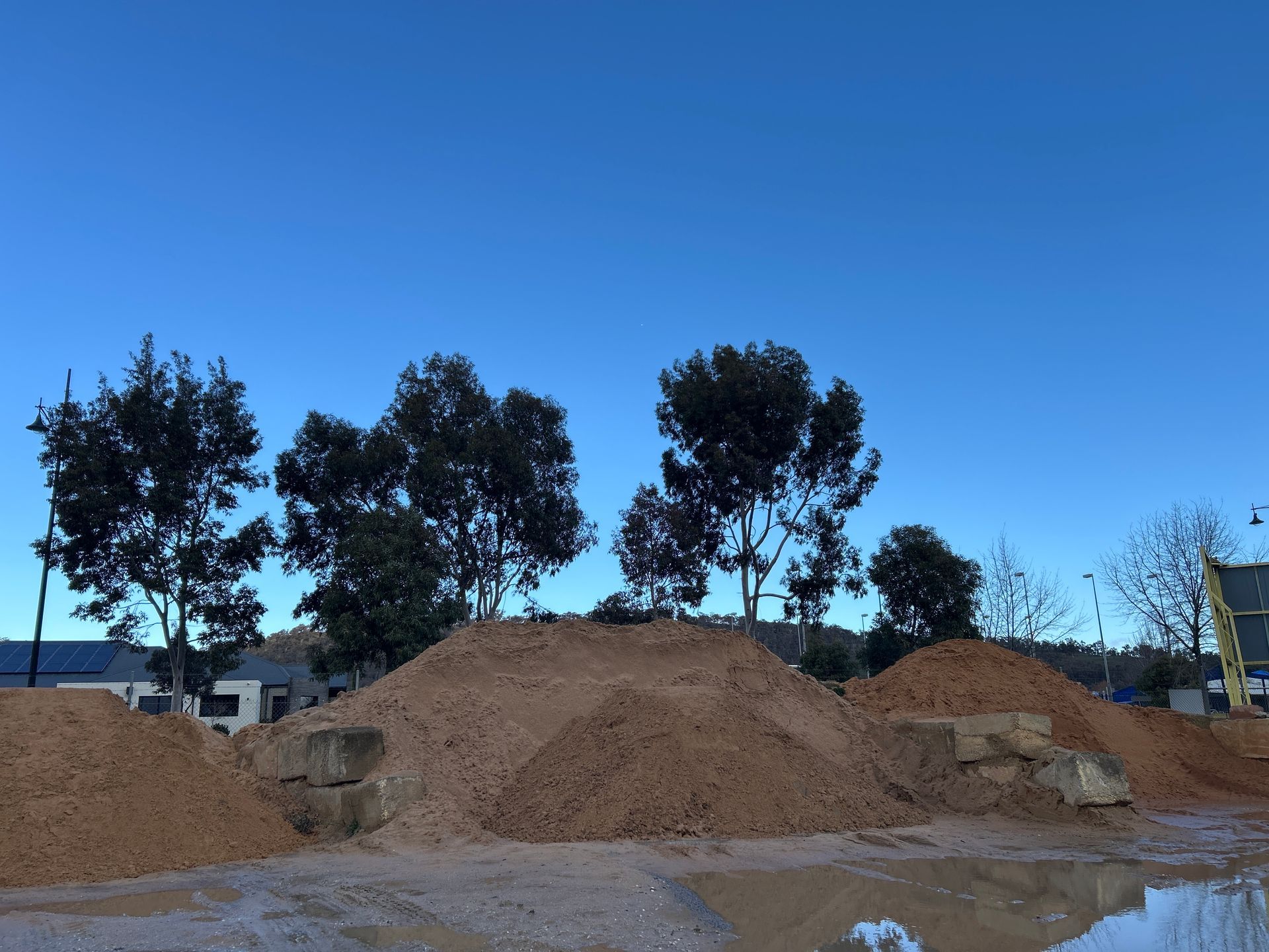 Piles Of Brown Material In Front Of Trees And Under A Clear Blue Sky. A Wet Surface Reflects Some Light — Wodonga Sand & Soil in Wodonga, VIC