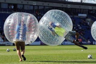 Two people in bubble suits colliding during a soccer game on a stadium field.