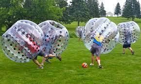 People playing bubble soccer on a grassy field, wearing large transparent inflatable spheres while chasing a ball.
