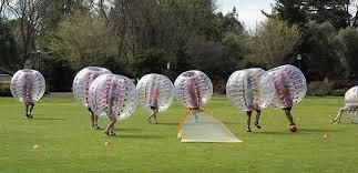 People in bubble suits play on a grassy field with a small goal net nearby.