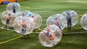 People playing bubble soccer on an indoor green turf field, wearing large transparent inflatable spheres.